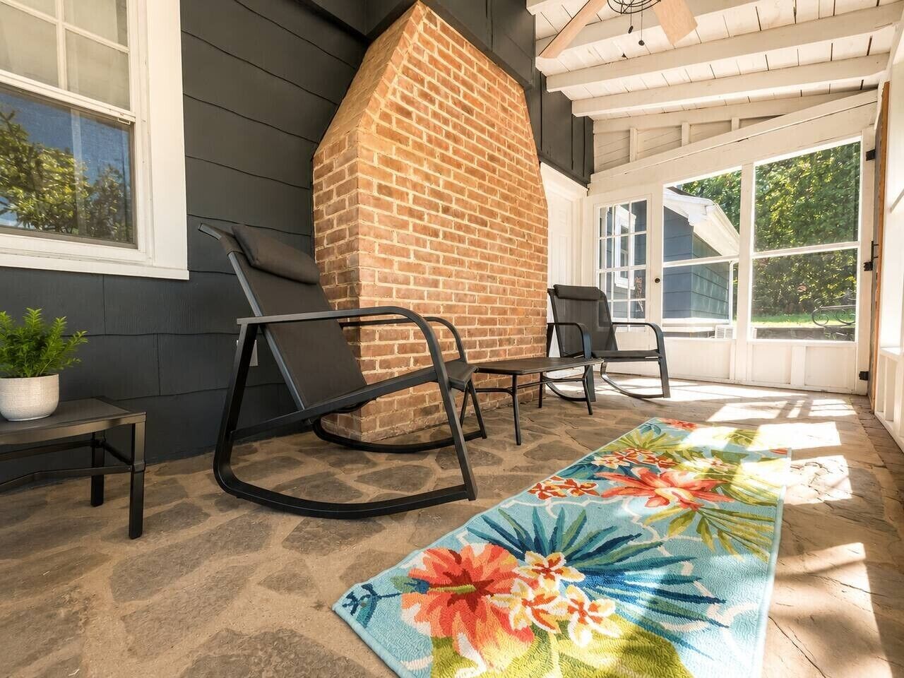 A covered porch with a brick fireplace, two black chairs, a floral rug, and a potted plant.