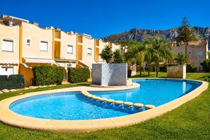 A swimming pool with a stone fountain, surrounded by a well-maintained lawn and a row of yellow buildings with balconies. - Ad3202 in Denia With 3 Bedrooms and 2 Bathrooms (Denia)