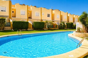 A swimming pool with a clear blue water surface, surrounded by a low hedge and a row of buildings with uniform yellow facades. - Ad3202 in Denia With 3 Bedrooms and 2 Bathrooms (Denia)
