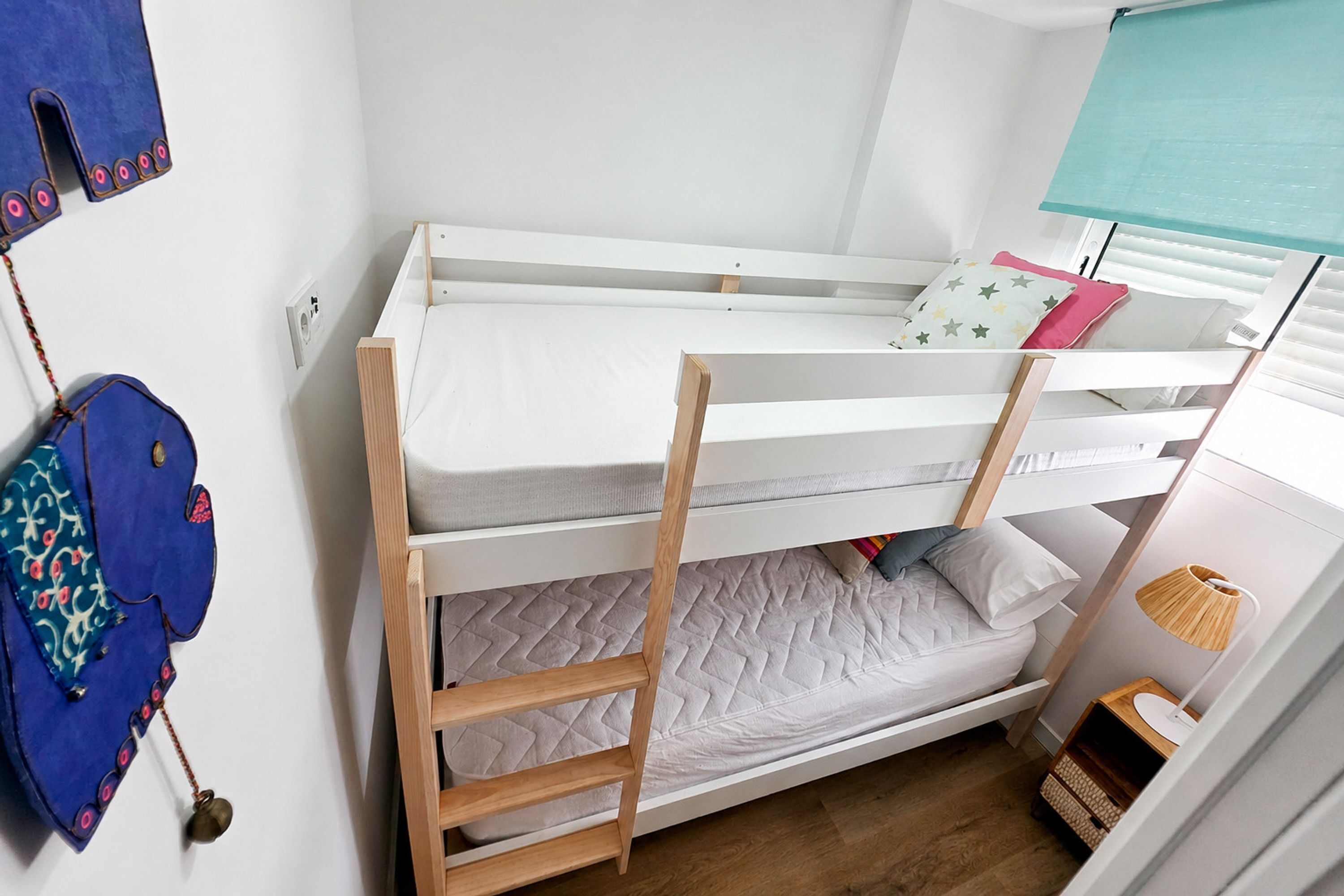 A bunk bed with a wooden ladder, a small bedside table with a lamp, and a wall-mounted bag.