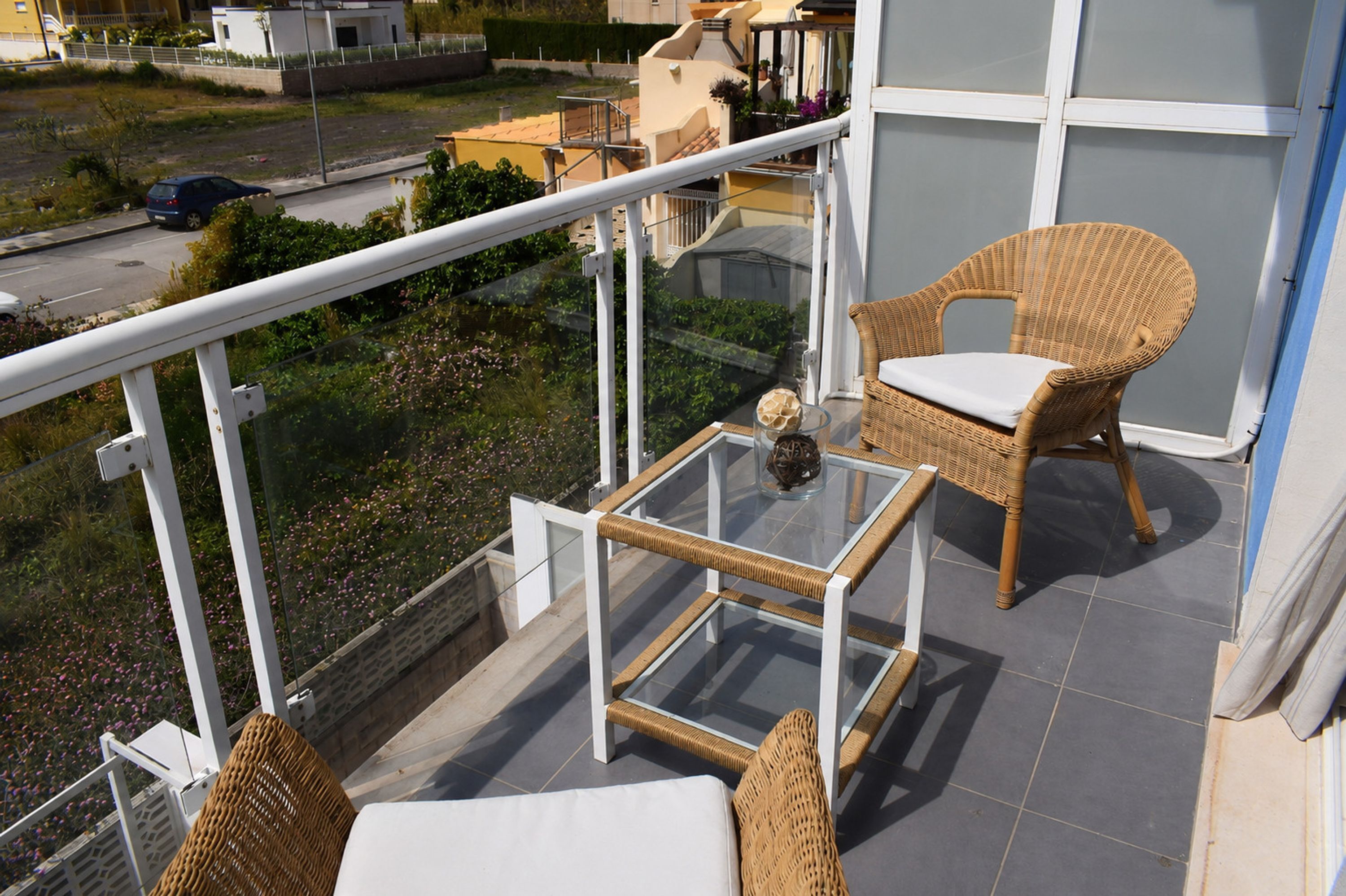 A balcony with wicker furniture, a glass table, and a view of the street and greenery.