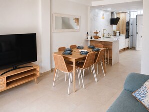 A modern living room with a flat-screen TV, a dining area with a wooden table and wicker chairs, and a kitchen with white cabinets and a built-in oven. - Ad1360 in Oliva With 3 Bedrooms and 1 Bathrooms (Oliva)
