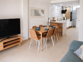 Una sala moderna con un televisor de pantalla plana, un comedor con mesa de madera y sillas de mimbre, y una cocina con alacenas blancas y horno empotrado.