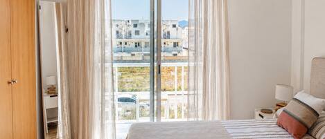 Un dormitorio con una cama grande, una mesita de noche, una lámpara, un armario y vistas a edificios por una ventana.