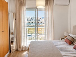 Un dormitorio con una cama grande, una mesita de noche, una lámpara, un armario y vistas a edificios por una ventana.