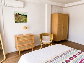 Un dormitorio con cama, cómoda de madera, silla de mimbre y armario.