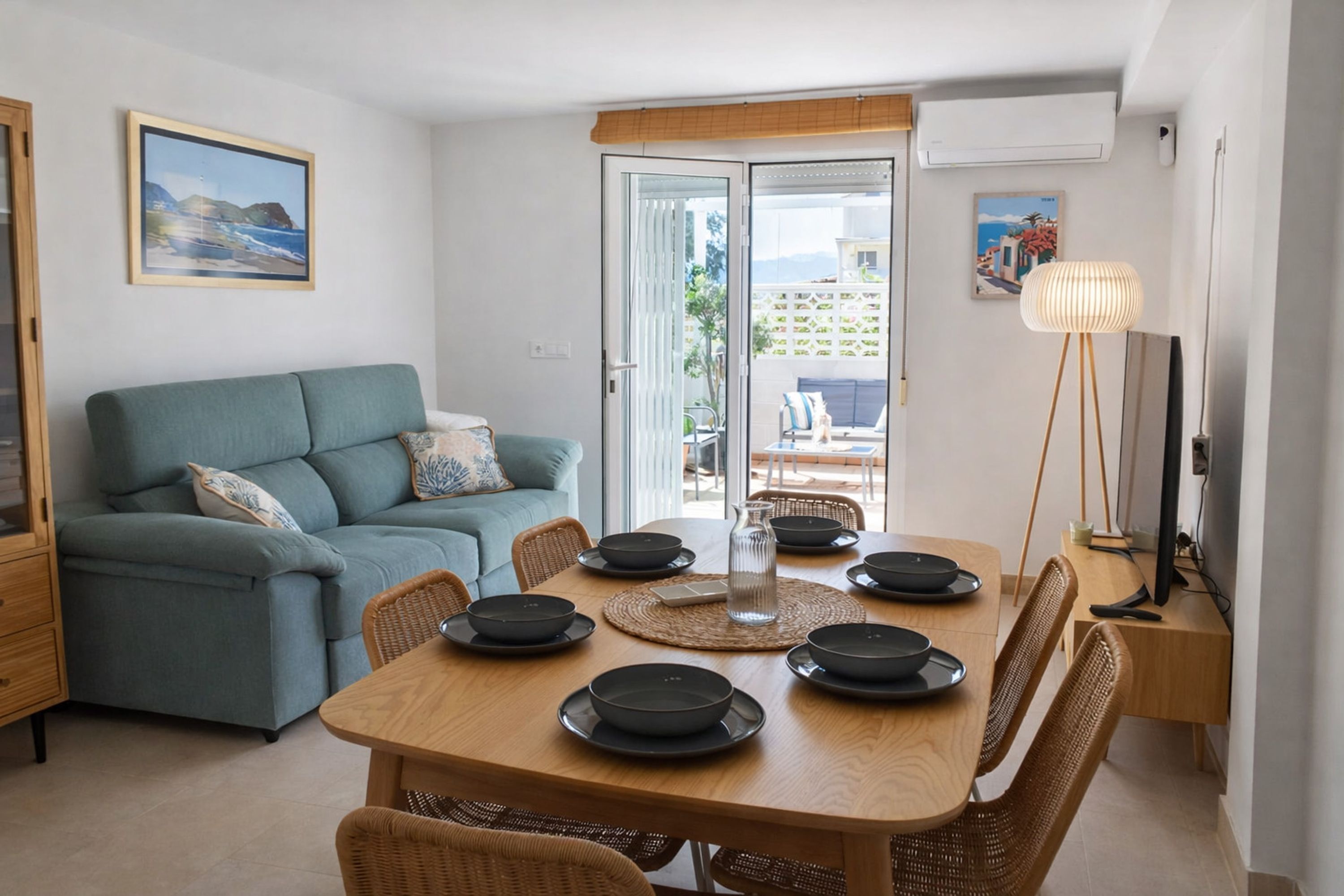 A dining area with a wooden table set for four, a teal sofa, wicker chairs, and a view through a glass door.