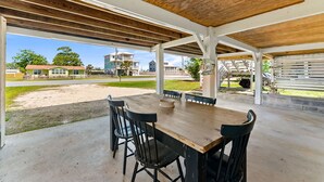 Um alpendre coberto ao ar livre com uma mesa e cadeiras de madeira, piso de concreto e vista para casas e vegetação ao fundo.