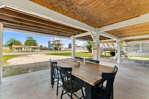 Eine überdachte Terrasse im Freien mit Holztisch und -stühlen, einem Betonboden und Blick auf Häuser und Grünpflanzen im Hintergrund.