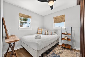Un dormitorio con cama, mesita de noche de madera, lámpara de pie, ventilador de techo, ventana con vistas y estante con objetos decorativos.