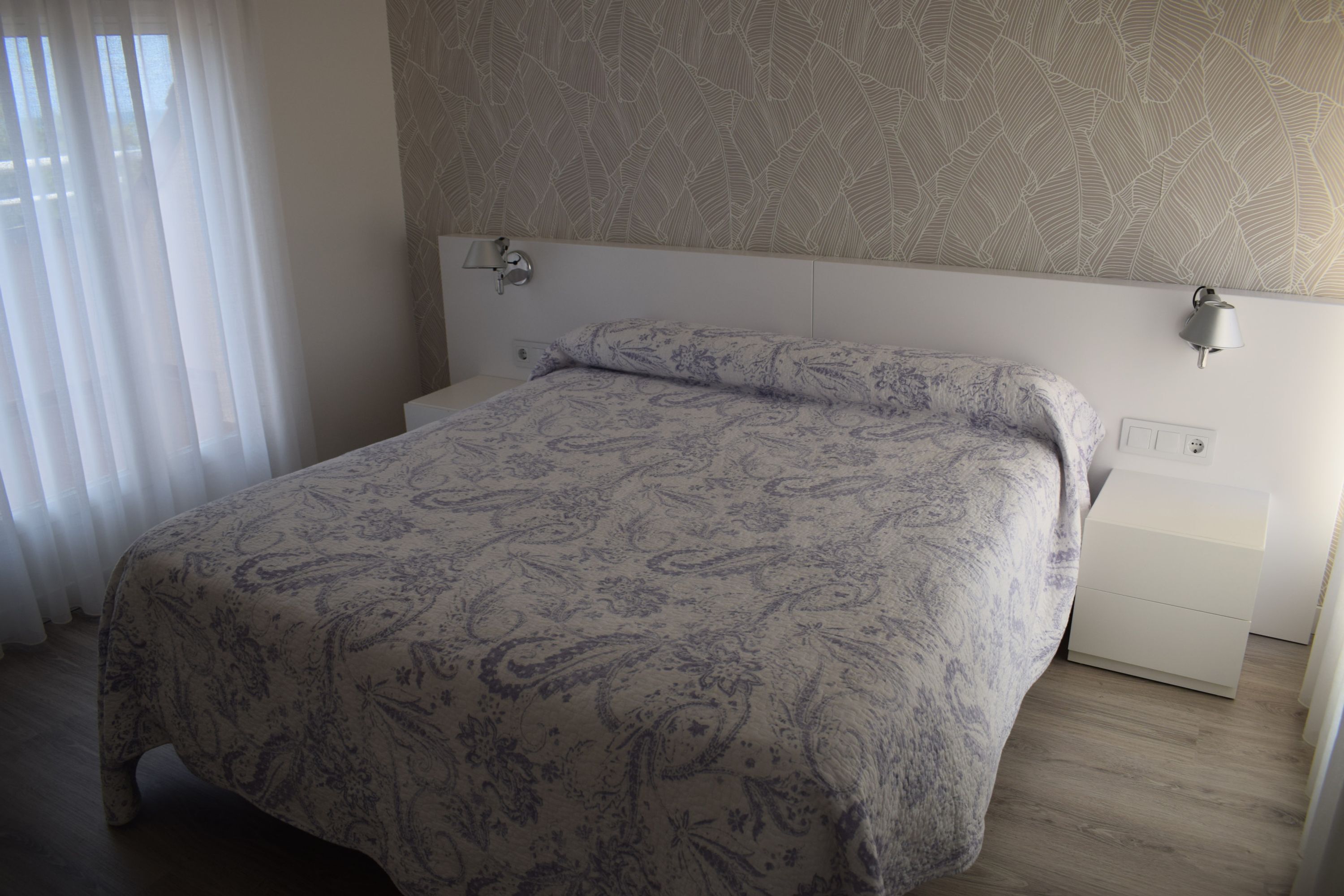 Een slaapkamer met een bed, nachtkastje, wandlamp en raam met gordijnen.