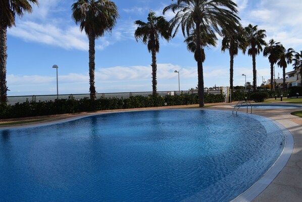 A swimming pool with a curved edge, surrounded by palm trees and a clear sky. - At2202 in Denia With 2 Bedrooms and 2 Bathrooms (Denia)