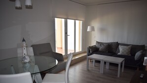 A modern living room with a glass dining table, white chairs, a grey sofa with cushions, and a floor lamp.