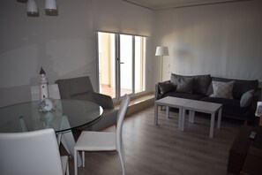 A modern living room with a glass dining table, white chairs, a grey sofa with cushions, and a floor lamp.