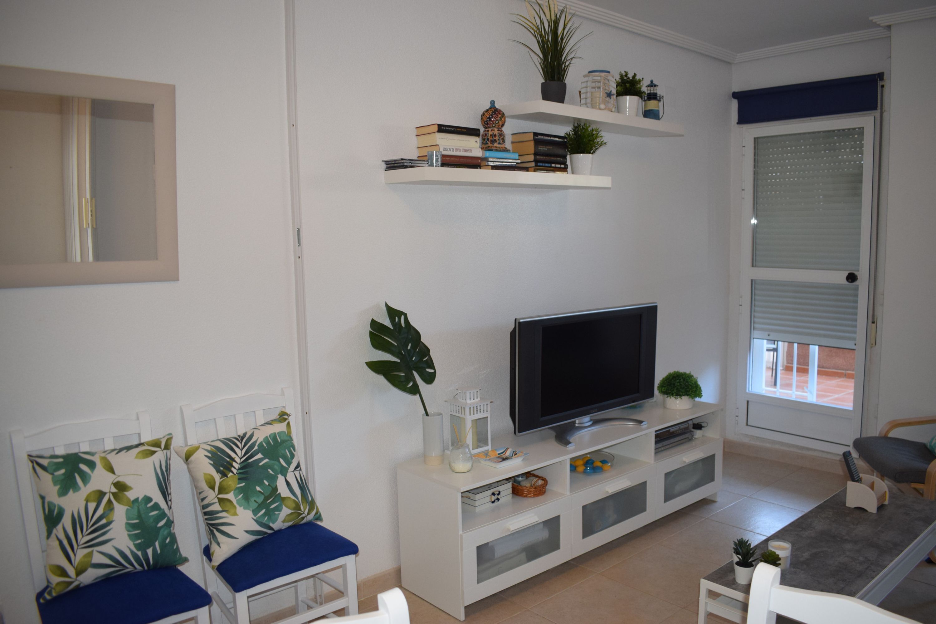 Una sala de estar con televisor de pantalla plana, muebles blancos, una estantería y un ventanal con persianas.