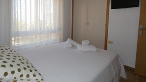 Un dormitorio con cama, armario, televisor y ventana con cortinas translúcidas.
