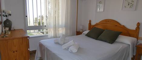 Una cama bien tendida con sábanas y toallas blancas, una mesita de noche de madera con una lámpara, un aparador de madera y una ventana con cortinas traslúcidas.