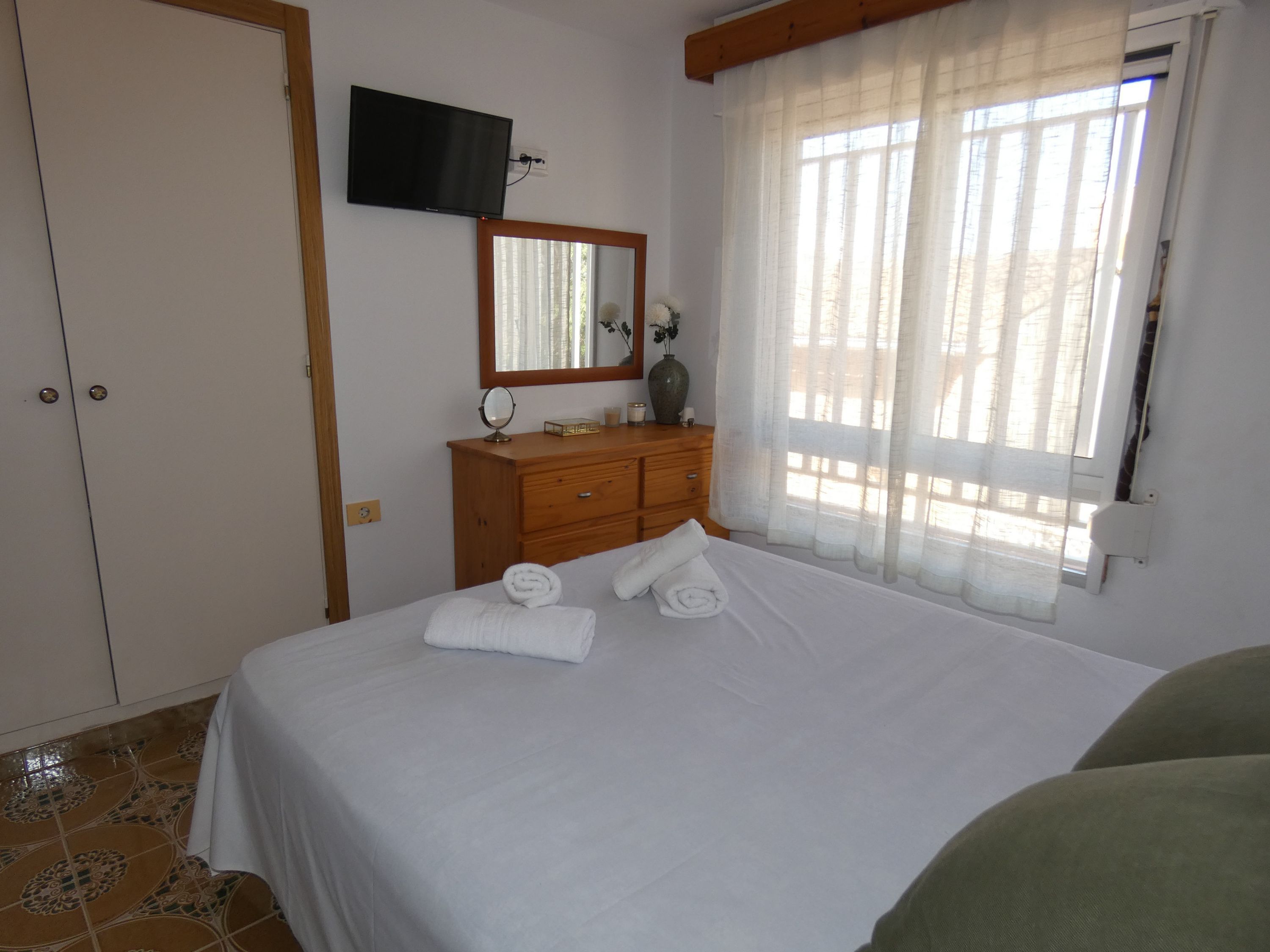 Un dormitorio con cama, cómoda, televisor montado en la pared y ventana con cortinas.