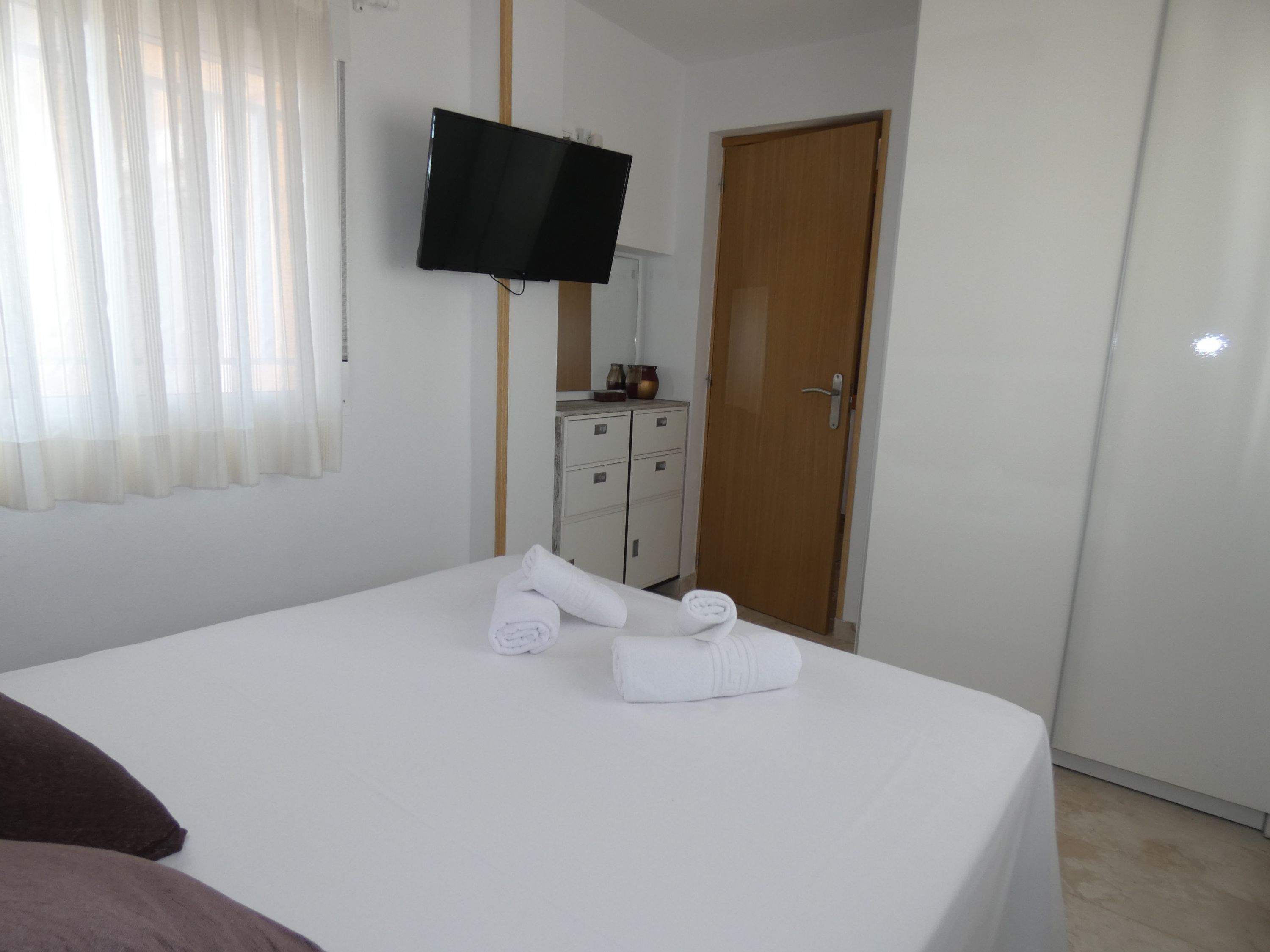 A hotel room with a bed, a television mounted on the wall, a wooden door, and a small cabinet.