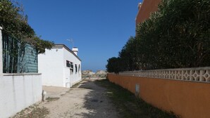 Un sendero de tierra estrecho entre edificios, uno de fachada blanca y otro de fachada naranja, que lleva a una colina distante.
