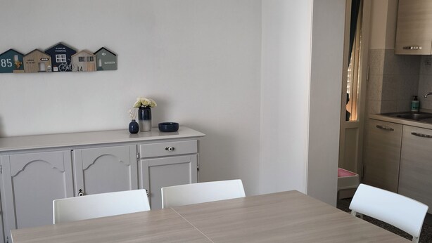 A dining area with a rectangular table and white chairs, a white cabinet with a shelf, and a wall-mounted decoration. - Vada Apartment - VadaLi (Vada)