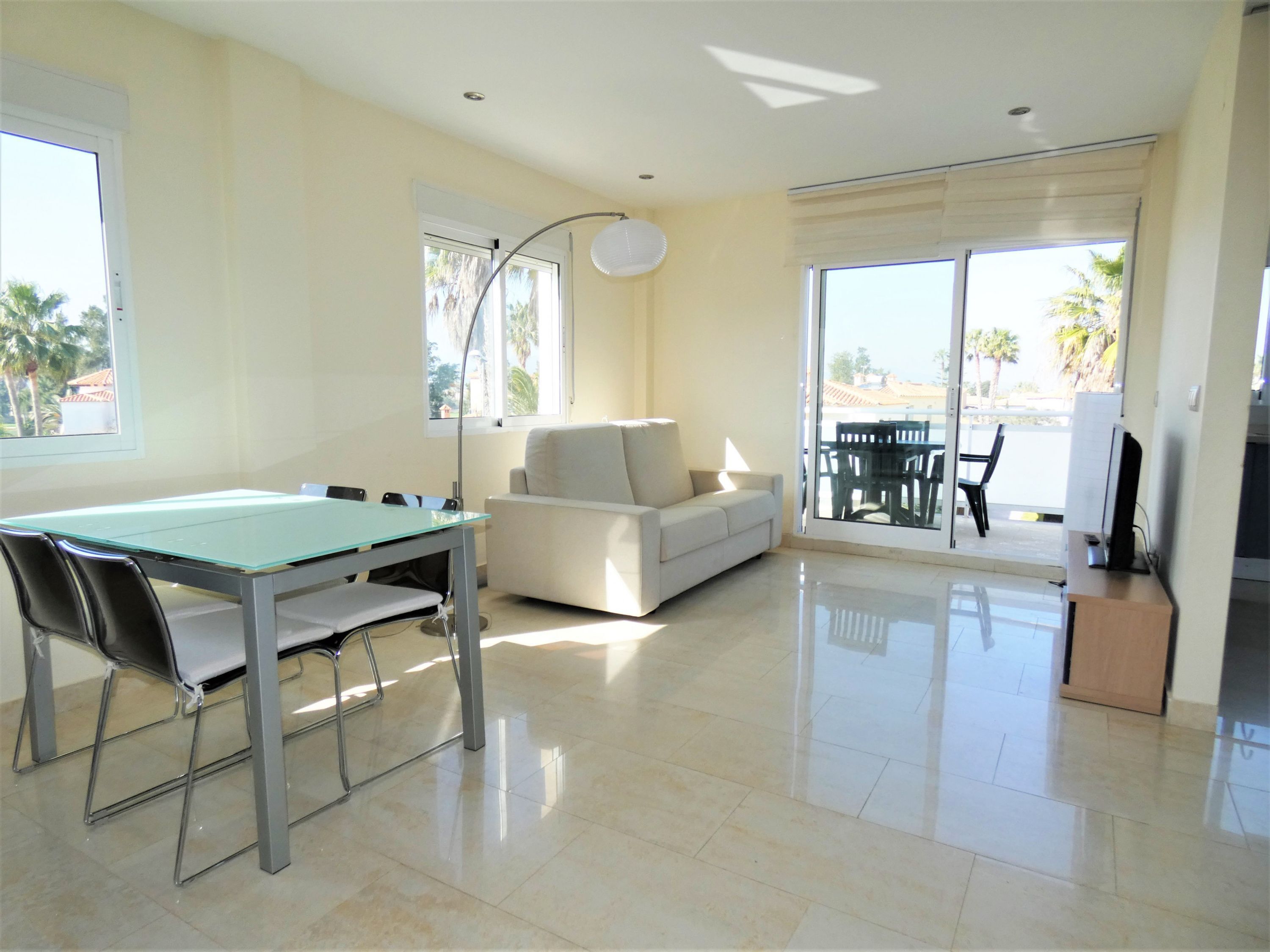 A spacious living area with a glass-top table, a sofa, and a balcony with outdoor furniture.