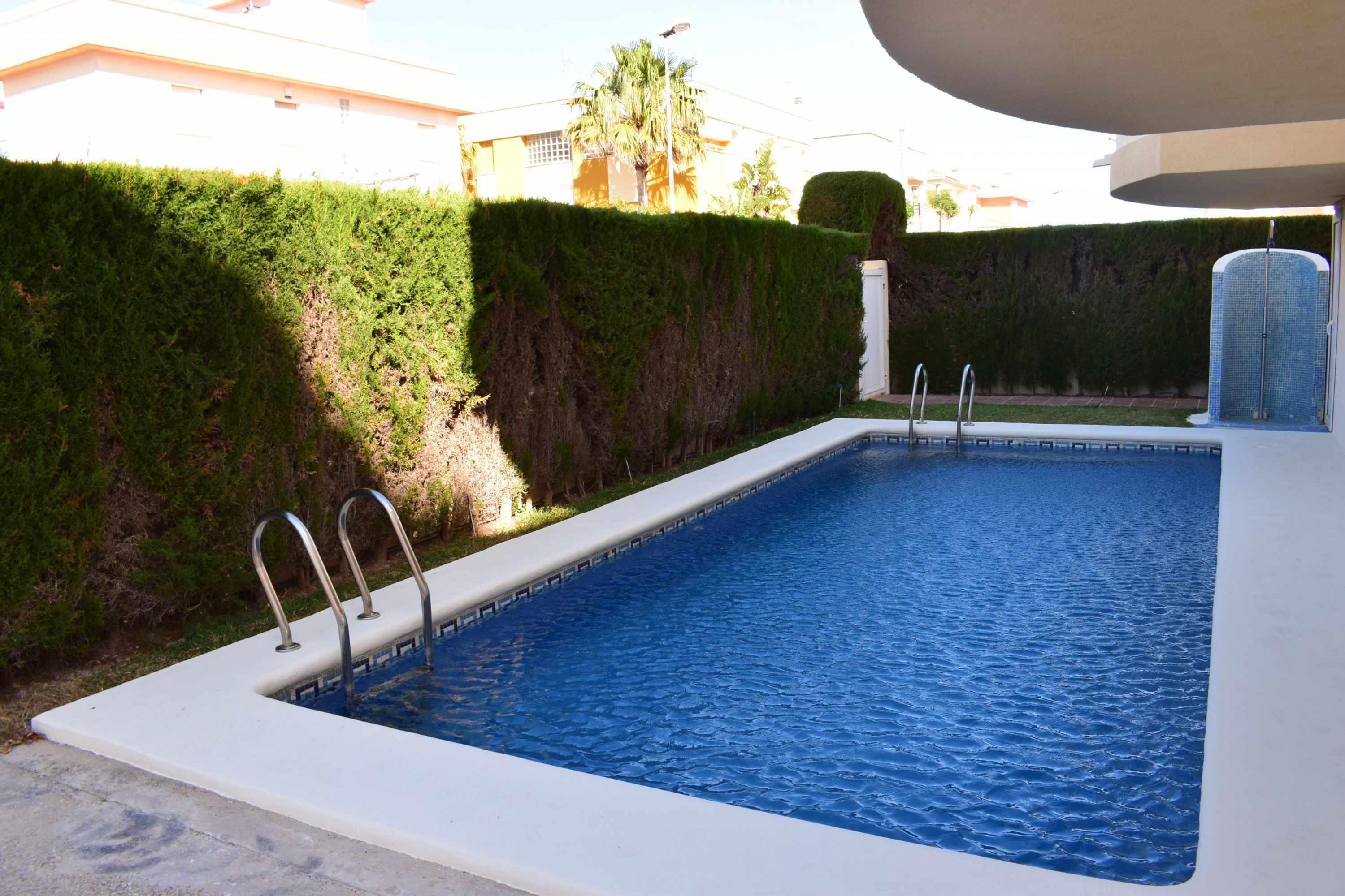 Uma piscina retangular com escadas de metal, cercada por um sebe baixo e um edifício ao fundo.
