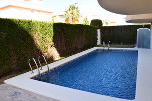 Una piscina rectangular con escaleras de metal, rodeada por setos bajos y un edificio al fondo.