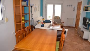 Uma sala de jantar com mesa e cadeiras de madeira, uma estante de livros, um sofá e uma televisão sobre um suporte.
