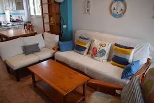Sala de estar con sofá, mesa de centro y mesita auxiliar de madera. Se ve una cocina al fondo.