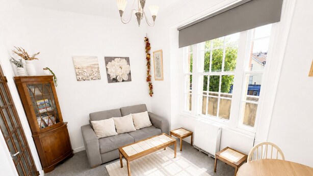 A living room with a grey sofa, a wooden coffee table, a small wooden table, a dining table, and a cabinet with glass doors. - Free Parking | Homely Gloucester Rd Flat Sleeps 3 (Bristol)