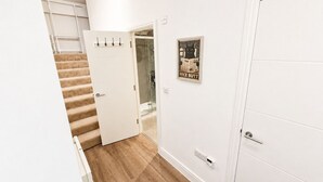 Um corredor com piso de madeira, porta branca, box de vidro e interruptor de luz na parede.
