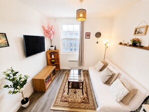 Ein Wohnzimmer mit einer Couch, einem Couchtisch, einem Fernseher, einem Bücherregal, einer Topfpflanze und einem Fenster.