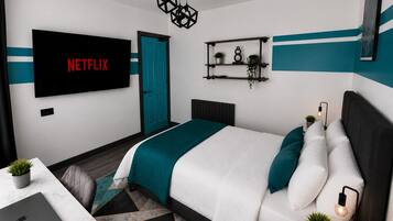 Une chambre moderne avec un lit, un bureau sur lequel se trouve un ordinateur portable, un téléviseur affichant le logo « NETFLIX », et une porte.