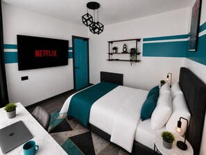 Un dormitori modern amb un llit, un escriptori amb un ordinador portàtil, un televisor que mostra 'NETFLIX' i una porta.