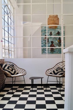 Ein Zimmer mit Schachbrettmuster-Boden, zwei Korbstühle, einem kleinen Tisch und einer Wand mit einem Fenster, das ein Bild von Menschen im Wasser zeigt.