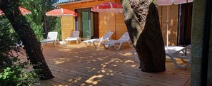 A wooden deck with white lounge chairs and red umbrellas, surrounded by trees.