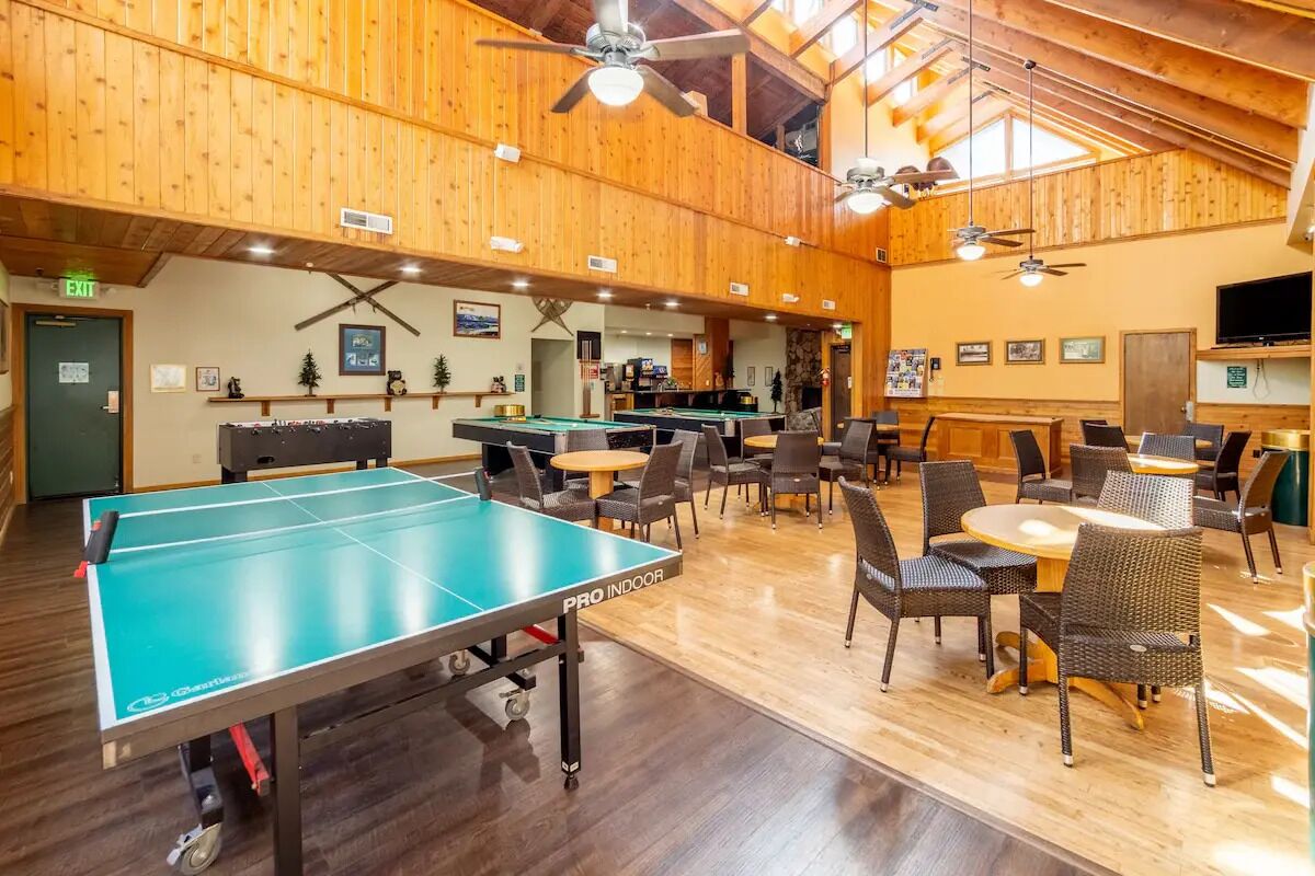 An indoor recreational area with ping pong tables, seating, and ceiling fans.