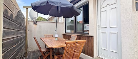 Un patio al aire libre con una mesa y sillas de madera, un paraguas negro y una puerta blanca con un número.