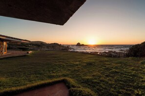 Paisagem costeira com um pôr do sol, terreno rochoso e uma área gramada.