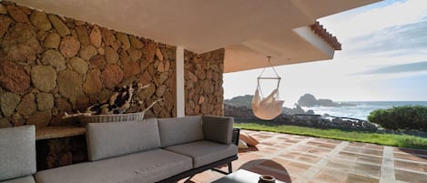 A stone wall, a sofa, a hanging chair, and a view of the ocean.