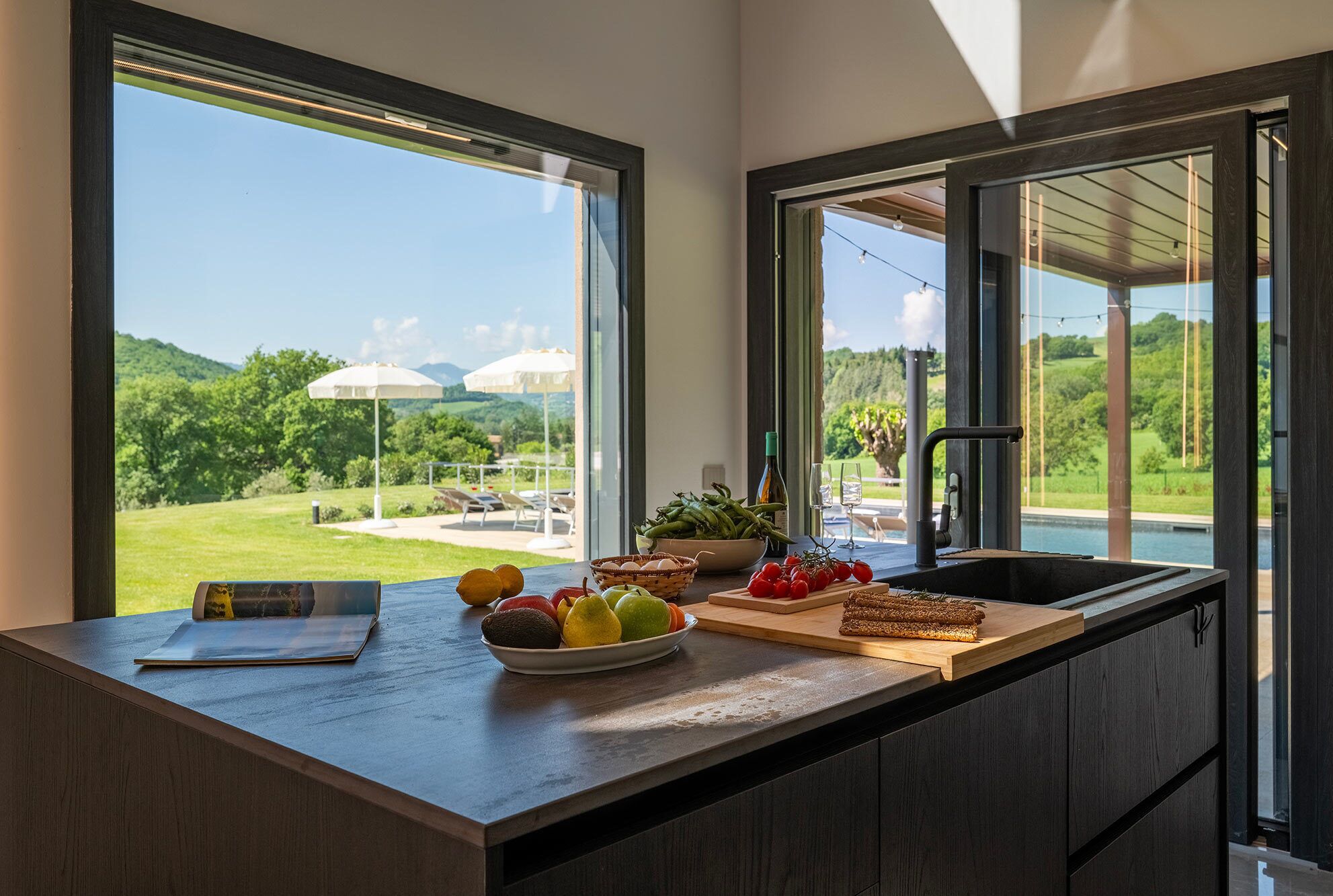 A kitchen with a large window, a bowl of fruit, a cutting board with vegetables, and a bottle of wine.