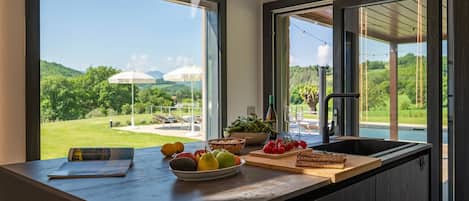 A kitchen with a large window, a bowl of fruit, a cutting board with vegetables, and a bottle of wine.