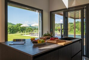 Eine Küche mit einem großen Fenster, einer Obstschüssel, einem Schneidebrett mit Gemüse und einer Weinflasche.