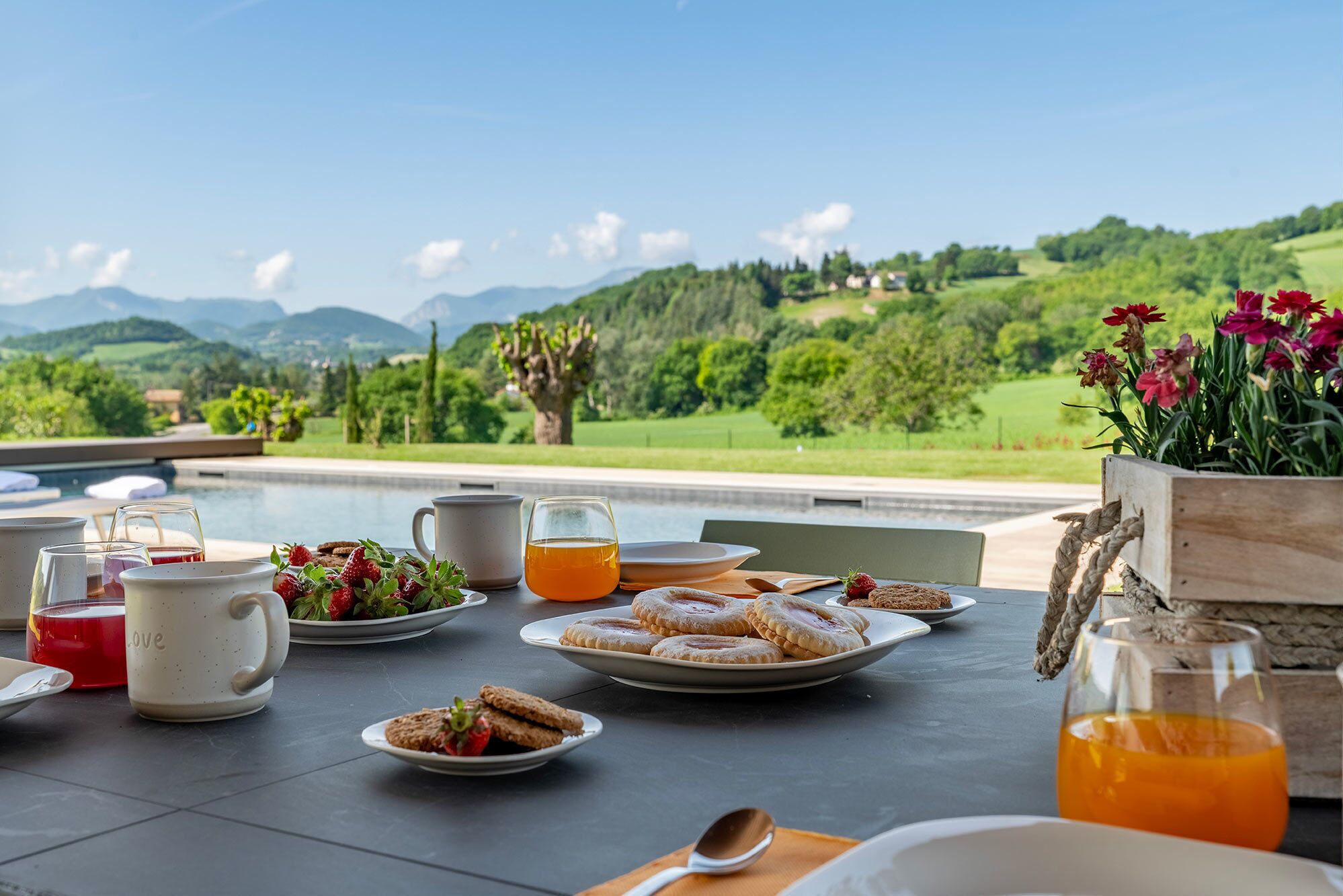 A breakfast table with plates of food, glasses of juice, and a view of a green landscape.