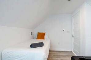 A small, white-walled room with a single bed, a door, and a small window.