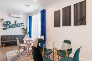 Ein modernes Wohnzimmer mit einer Couch, einem Glastisch mit Stühlen, einem blauen Vorhang und einer Wand-Klimaanlage.