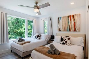 Une chambre d’hôtel avec deux lits, un ventilateur de plafond, un tableau et de grandes fenêtres donnant sur un paysage verdoyant.