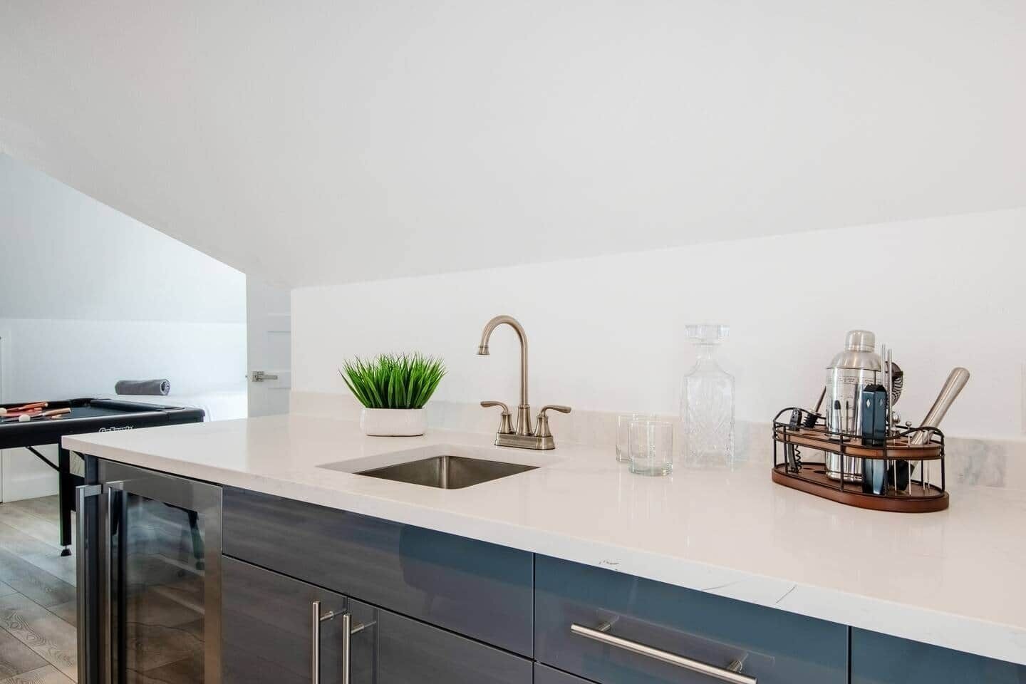 A modern kitchen with a sink, a potted plant, and a bar area with glasses and a cocktail shaker.