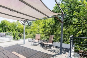 Eine überdachte Terrasse im Freien mit zwei Stühlen und einem Tisch, umgeben von einem Metallgeländer und Bäumen.